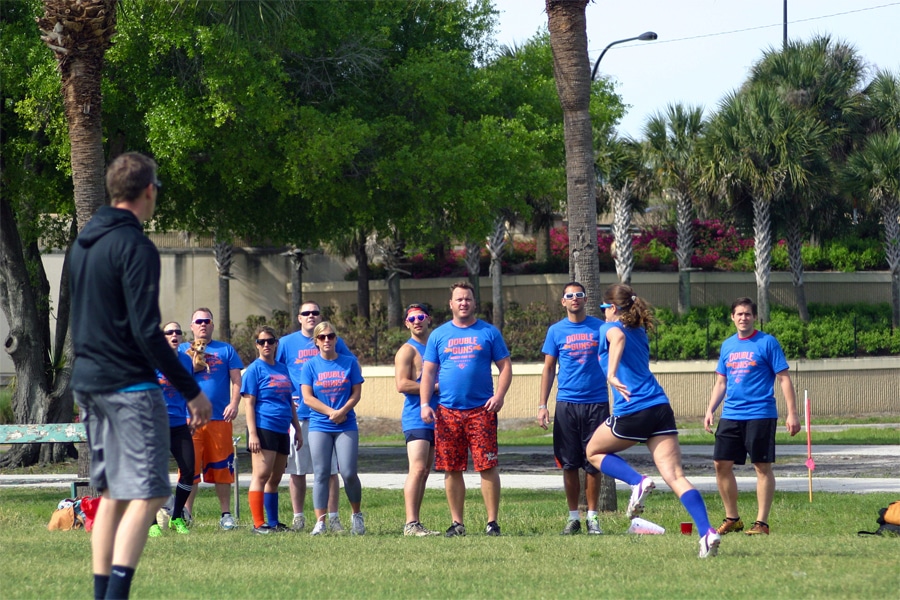  group of people wearing matching blue “Double Guns” shirts watch a woman in athletic wear running and preparing to kick a ball on a grassy field.