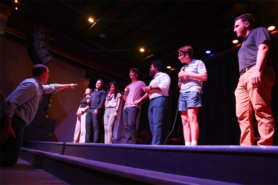A group of students stand on stage holding microphones, engaged in an interactive workshop led by a kneeling instructor pointing toward them.