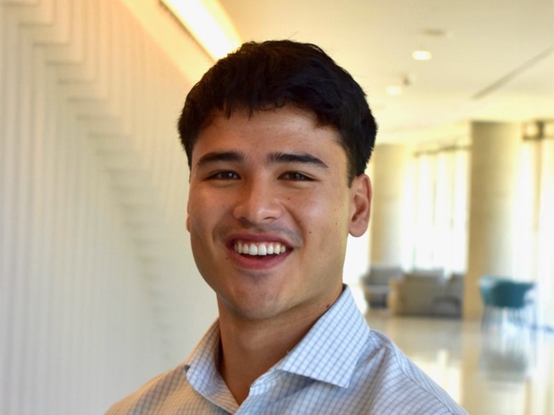 A young man in a light-colored button-down shirt smiles warmly at the camera in a bright, modern hallway.