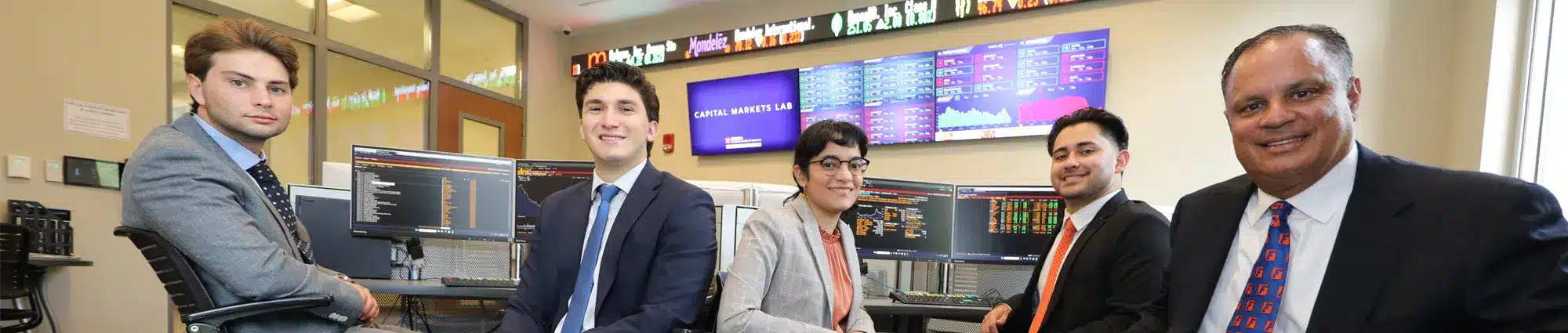 Five finance students and a professor gather around a computer setup in the Capital Markets Lab, reviewing investment data on multiple screens.