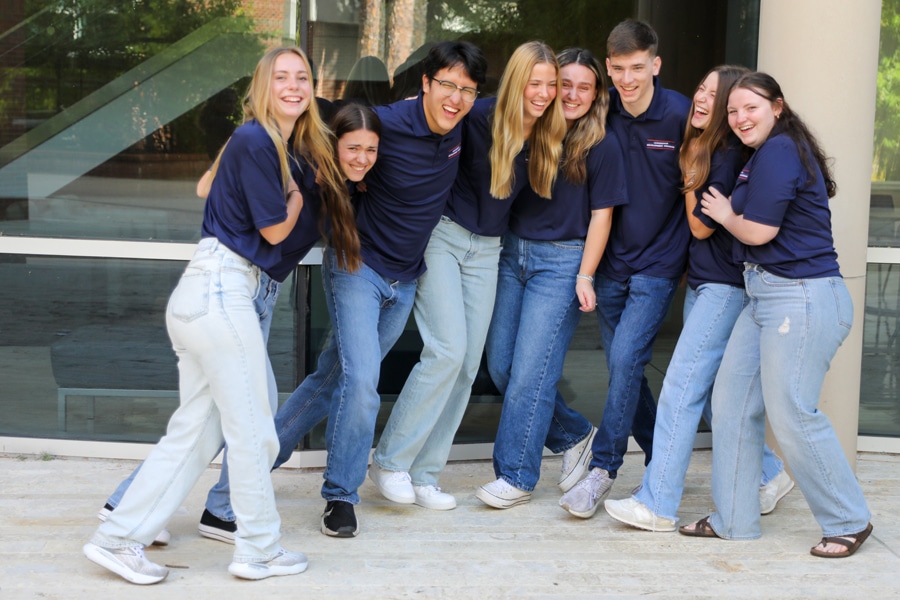 A group of eight students in matching navy polo shirts and jeans stand outside, smiling and laughing as they pose closely together for a team photo.