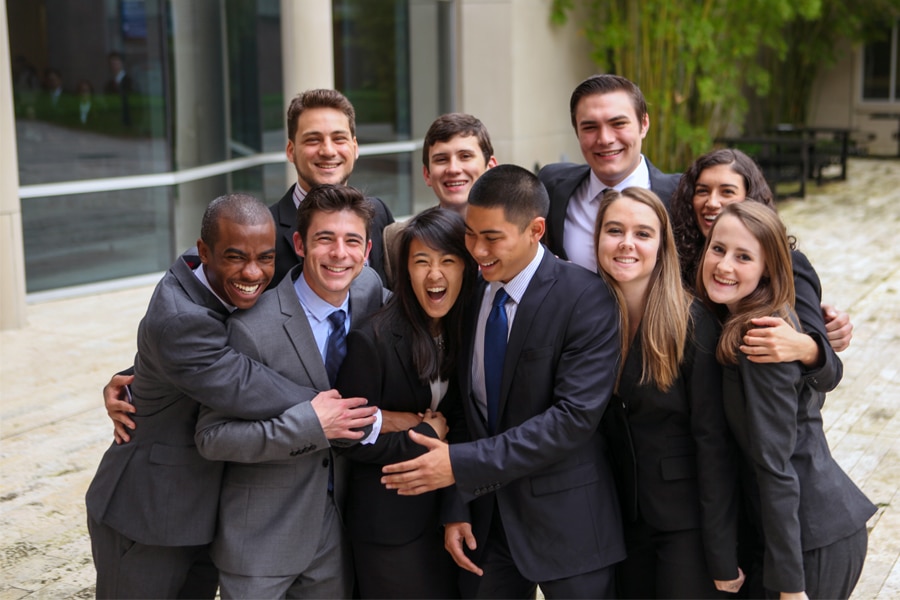 diverse group of ten University of Florida business students, dressed in professional attire, standing outside near a building. They are grouped tightly together in a joyful, warm embrace, smiling and laughing.