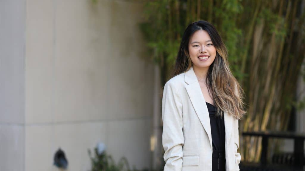 A confident young woman in professional attire smiles outdoors near campus.