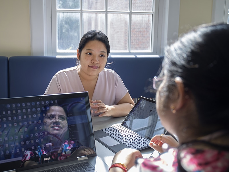 Two students sitting at a table with laptops open, engaged in a discussion. One student’s face is visible, looking attentively at the other, whose reflection appears on the laptop screen.
