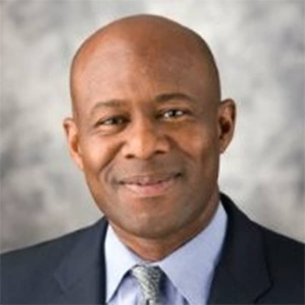 Professional headshot of Algernon Callier, a bald Black man wearing a dark suit jacket, light blue shirt, and patterned tie, smiling gently with a gray blurred background.