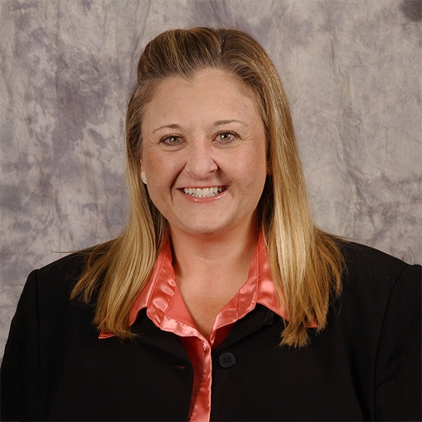 Professional portrait of Andria Long, a woman with long blonde hair, smiling, wearing a black blazer over a shiny coral blouse, against a gray mottled backdrop.
