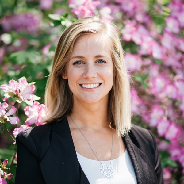 Portrait of Ashley Turner, smiling, with straight blonde hair, wearing a black blazer and white top, standing in front of a background of vibrant pink flowers.
