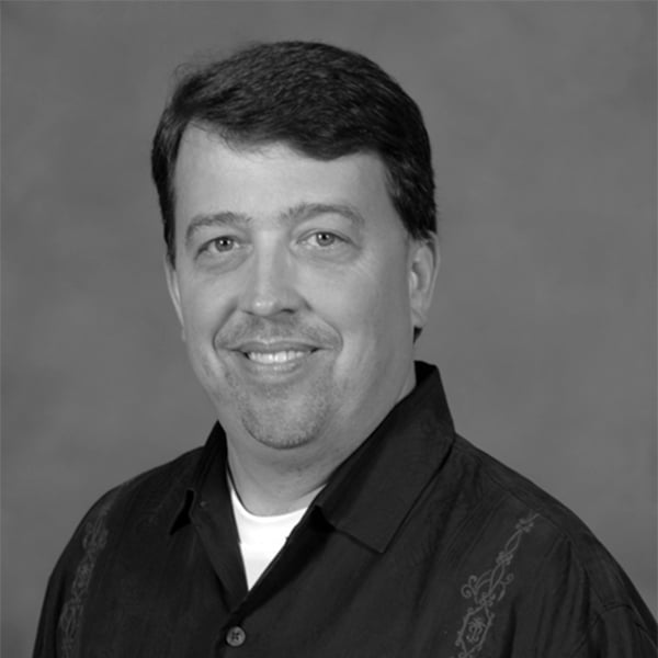 Black and white professional headshot of Brent Berthy, a man with short dark hair, a goatee, and a warm smile, wearing a dark patterned button-up shirt.