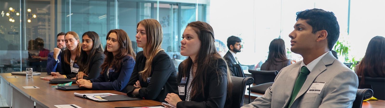 A group of students in professional attire attentively listen during a meeting in a modern conference room with large windows.