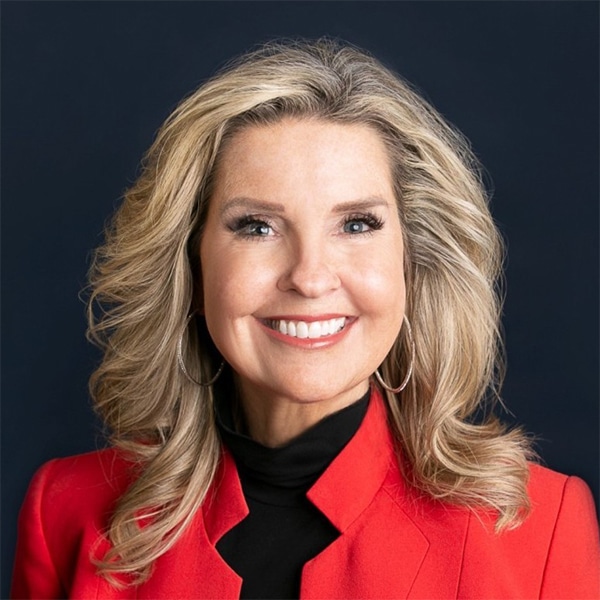 Professional headshot of Cheryl Cook, a blonde woman with shoulder-length hair, wearing a bright red blazer over a black top, smiling confidently against a dark blue background.