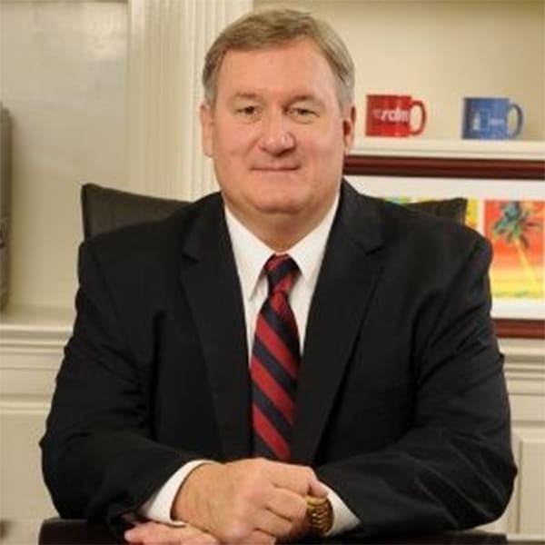 Professional portrait of Chip Lane, a middle-aged man with light brown hair, wearing a black suit, white shirt, and a red-and-blue striped tie, seated with hands clasped on a desk, with a bookshelf and colorful mugs in the background.