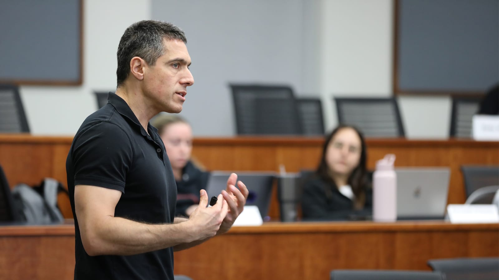 A professor speaks passionately at the front of a classroom while Ph.D. students listen in the background.