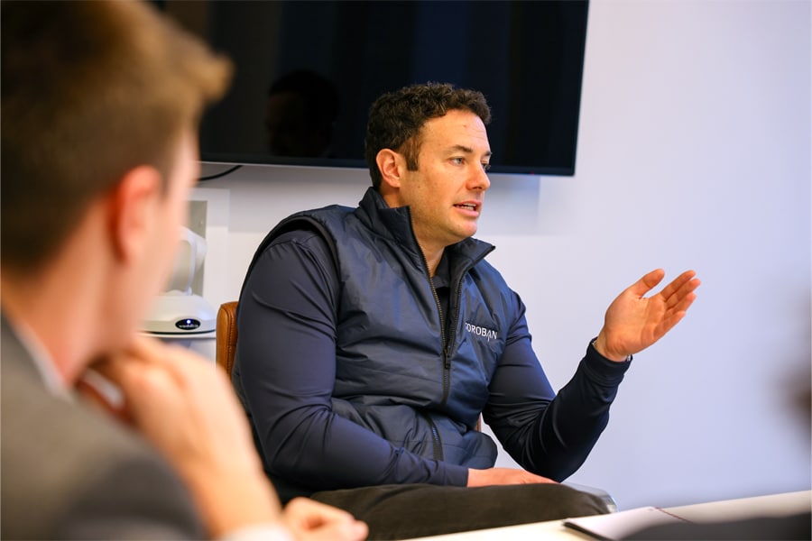 A business professional in a navy vest and shirt gestures as he speaks during a small group discussion in a modern conference room.