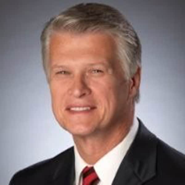 Professional headshot of Dan Doyle, smiling slightly, with short silver hair, wearing a dark suit, white shirt, and red striped tie, against a soft gray background.
