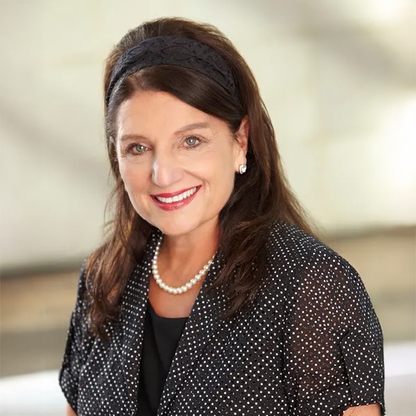 Headshot of Debbie Ferree, a middle-aged woman with light skin and long brown hair, wearing a black blouse with white polka dots, pearl necklace, and a black headband, smiling warmly against a blurred neutral background.