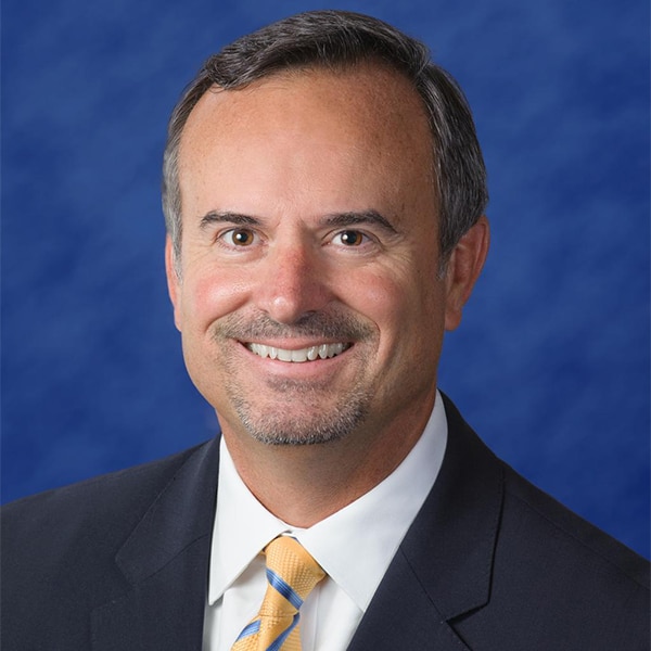 Professional headshot of Drew Asher, a man with short dark hair and a trimmed goatee, wearing a dark suit, white shirt, and yellow and blue striped tie, smiling against a blue background.