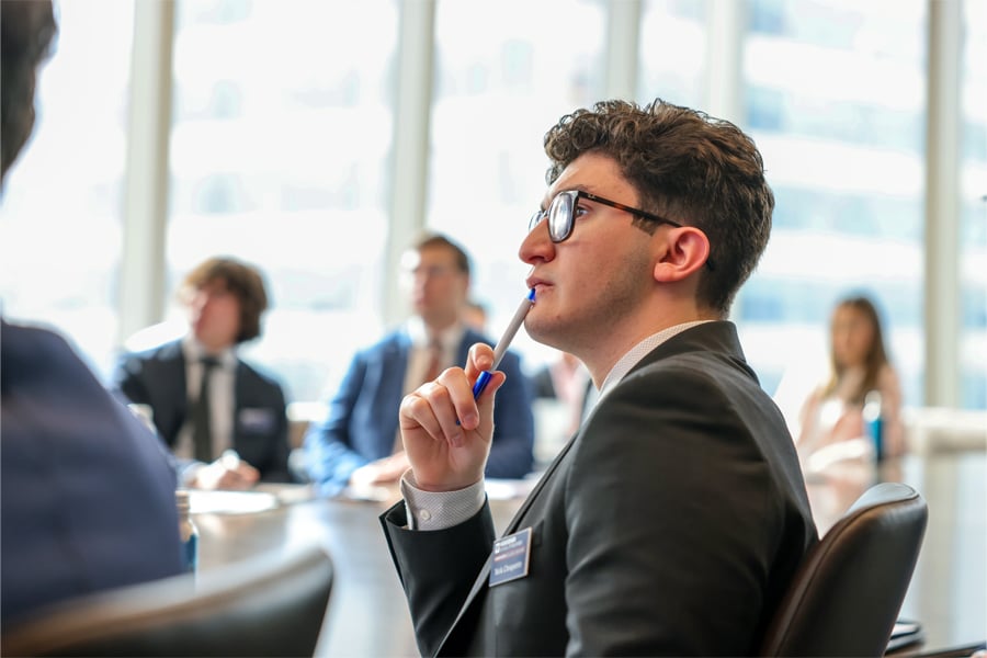 A focused young man in a business suit and glasses holds a pen to his lips thoughtfully during a group meeting, with blurred colleagues seated around a conference table.