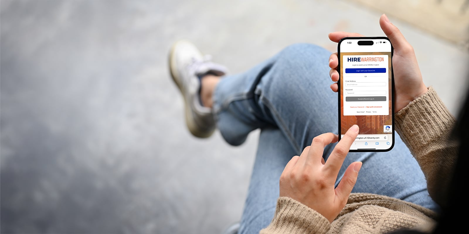 A student's hands holding a mobile device displaying the HIREWarrington login page. 