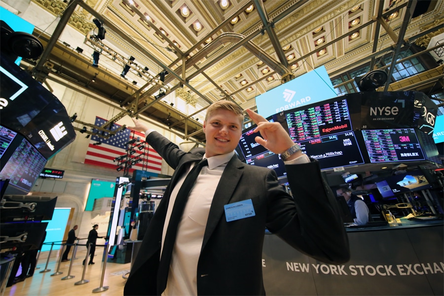 Young man in a black suit and white shirt poses excitedly on the floor of the New York Stock Exchange, with digital stock ticker screens and American flag visible in the background.