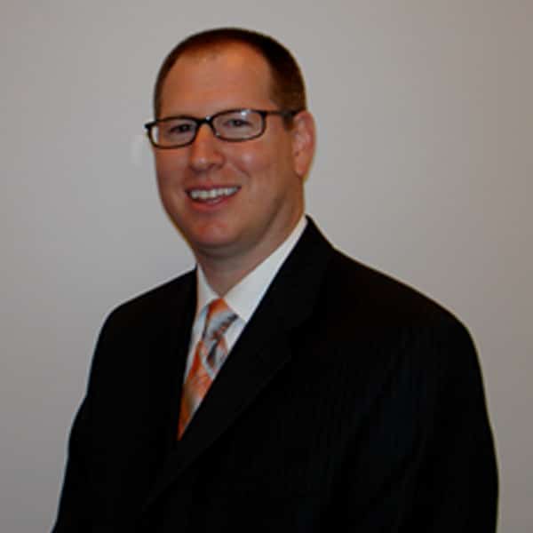 Headshot of James Lewis, smiling, wearing glasses, a dark pinstripe suit, white shirt, and an orange patterned tie, against a plain beige background.