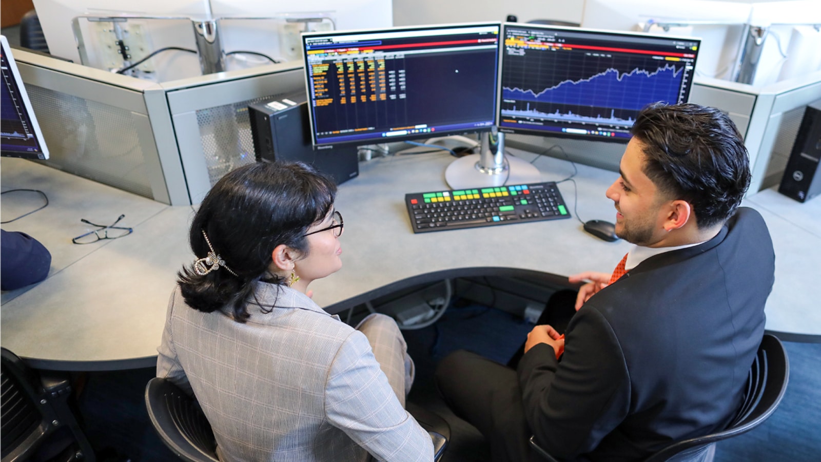 Two students in professional attire discuss investment data displayed on dual computer monitors in a modern trading room.