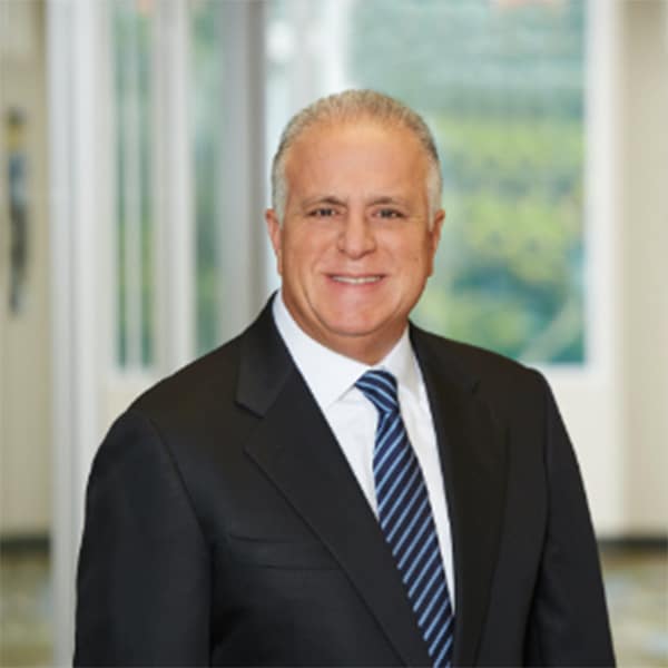 Professional headshot of Jorge Benitez, a man with gray hair wearing a black suit, white shirt, and a striped blue tie, smiling with a blurred window and greenery background.