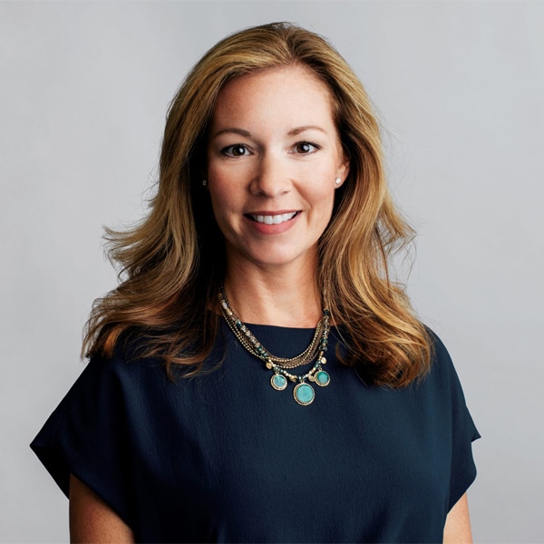 Portrait of Katherine Saez, smiling, with shoulder-length light brown hair, wearing a dark navy blouse and a layered turquoise and gold necklace, against a light gray background.