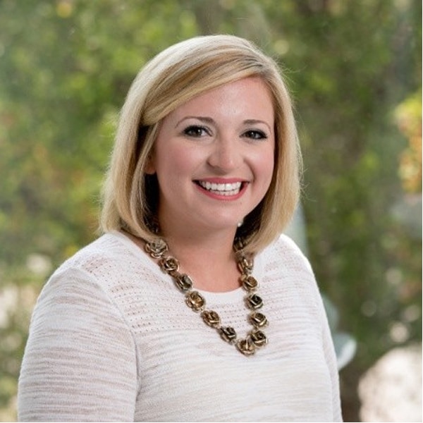 Portrait of Kayley McCranie, smiling, with straight blonde hair, wearing a white textured top and a chunky gold necklace, in front of a softly blurred natural background.