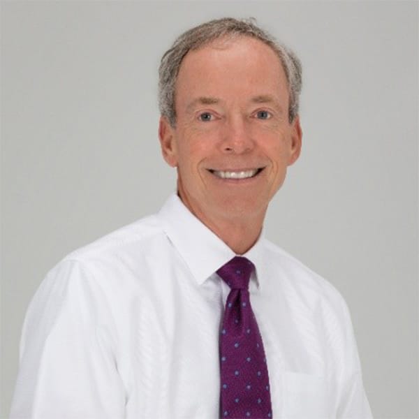 Professional headshot of Keith Koenig, a man with short gray hair, smiling, wearing a white dress shirt and a purple tie with blue dots, against a plain light gray background.