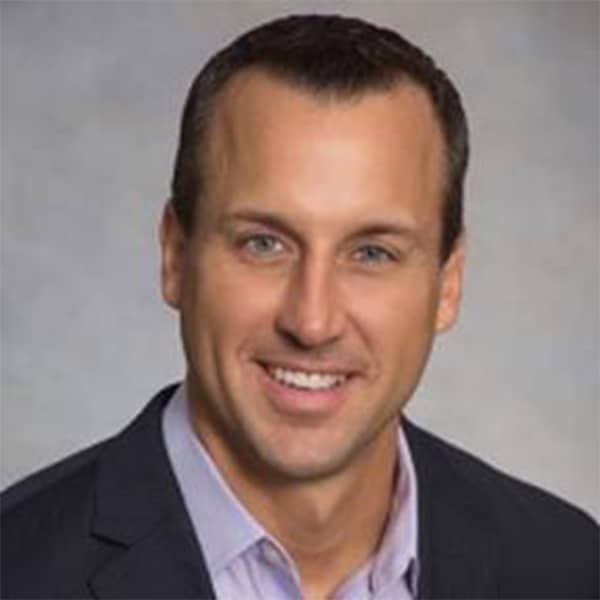 Professional headshot of Steven Kiker, a man with short brown hair and blue eyes, smiling, wearing a dark suit jacket and a light blue dress shirt, against a gray background.