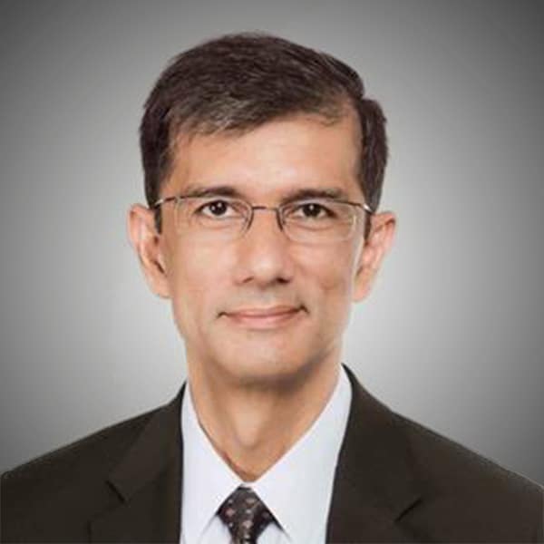 Professional headshot of Anjan Lahiri, a man with short dark hair and glasses, wearing a black suit, white shirt, and a patterned tie, against a light gray background.