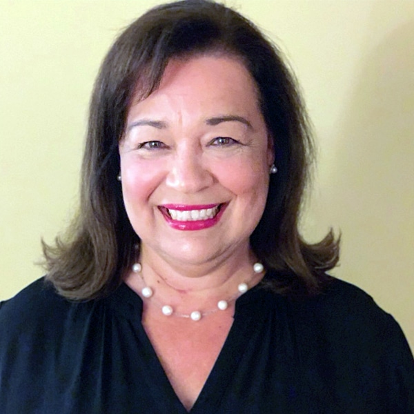 Headshot of Laura Guggenheim, a woman with shoulder-length dark brown hair, smiling warmly, wearing a black top and a pearl necklace, against a light yellow background.
