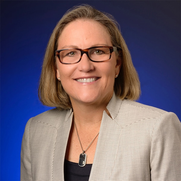 Professional headshot of Laurie Burns, a woman with shoulder-length light brown hair, wearing glasses and a beige blazer over a dark top, smiling warmly against a blue gradient background.