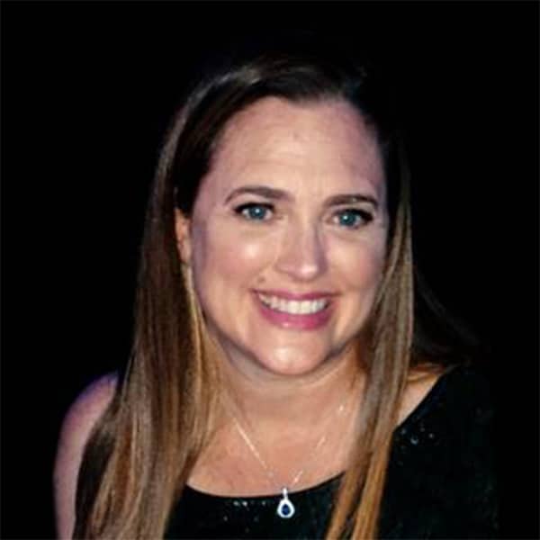 Headshot of Lorie Gleim, a woman with long light brown hair, blue eyes, and a bright smile, wearing a black top and a pendant necklace, against a dark background.