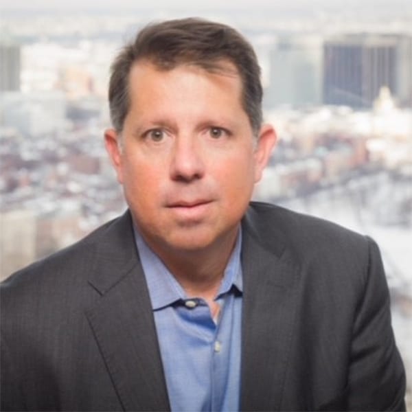 Professional portrait of Joe Marconi, a man with short brown hair and brown eyes, wearing a dark gray blazer and a blue button-up shirt, with an urban cityscape blurred in the background.