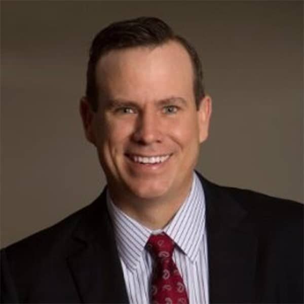 Professional portrait of Chris Meyer, a man with short brown hair, smiling, wearing a black suit jacket, light blue striped shirt, and red patterned tie, against a brown background.