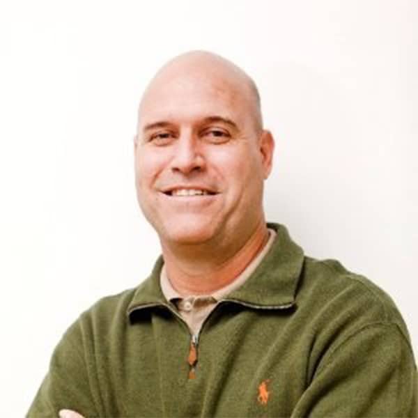 Professional portrait of Tony Milikin, a bald man smiling warmly, wearing a green quarter-zip fleece jacket over a beige shirt, standing against a plain white background.