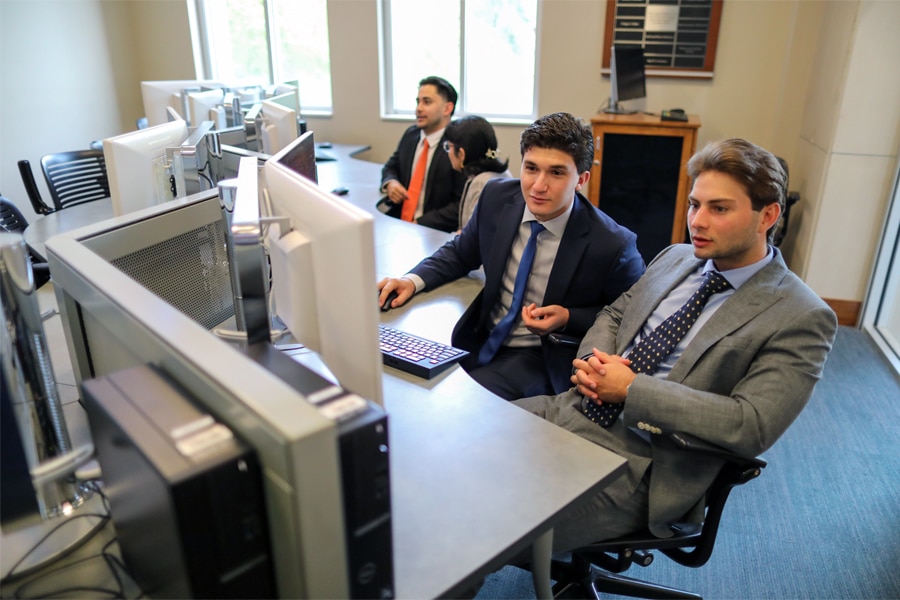 Two young men in suits intently watch multiple computer screens in a bright office, while two others work in the background.