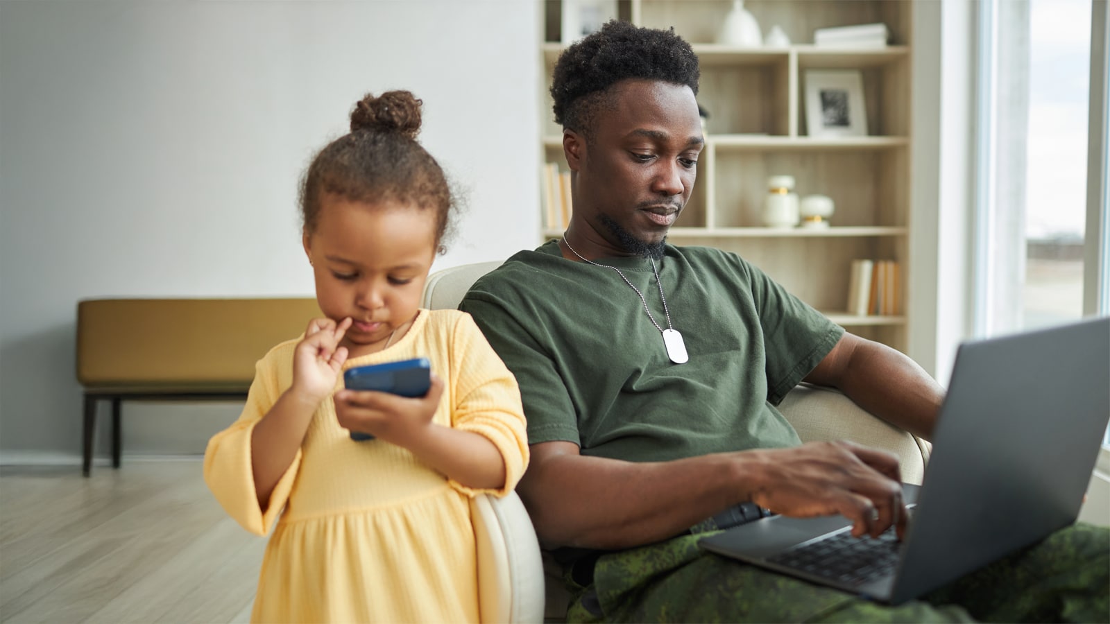 Military father works on laptop at home beside his young daughter