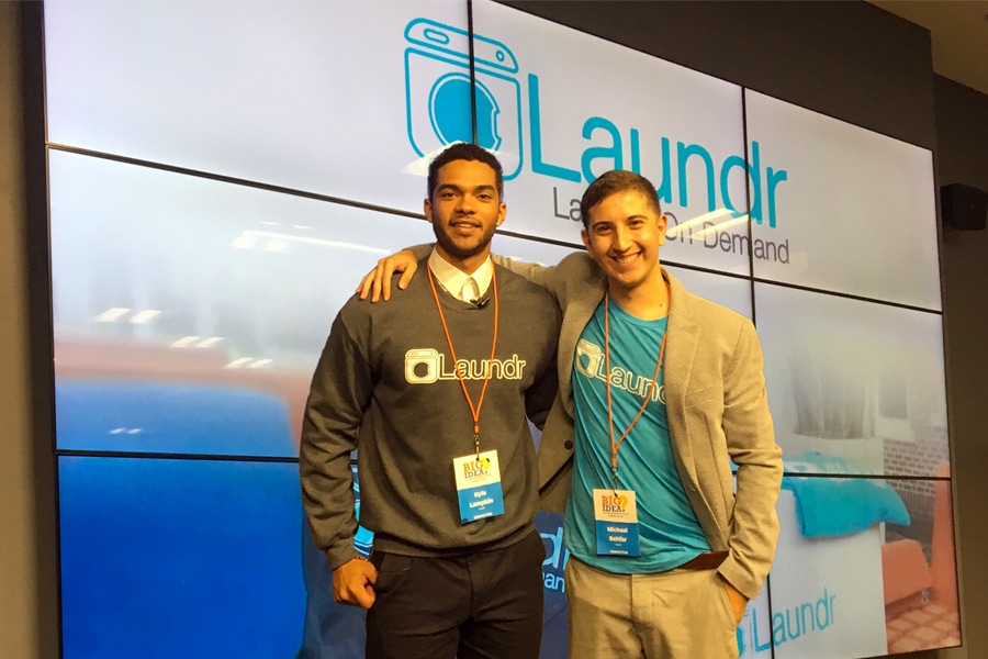 Two student entrepreneurs stand in front of a screen displaying their startup logo after presenting their business idea.