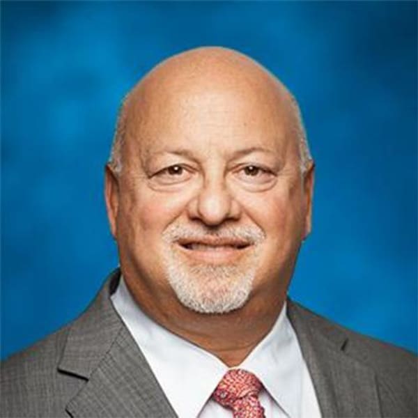 Professional headshot of Ben Plotkin, a middle-aged man with a bald head and white beard, wearing a gray suit, white shirt, and pink tie, smiling against a blue background.