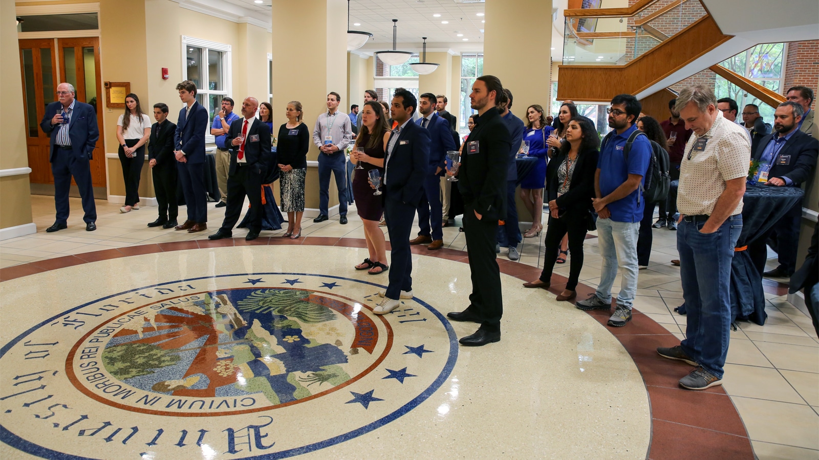 A diverse group of students and professionals stand attentively in a campus building, gathered around the University of Florida seal on the floor.