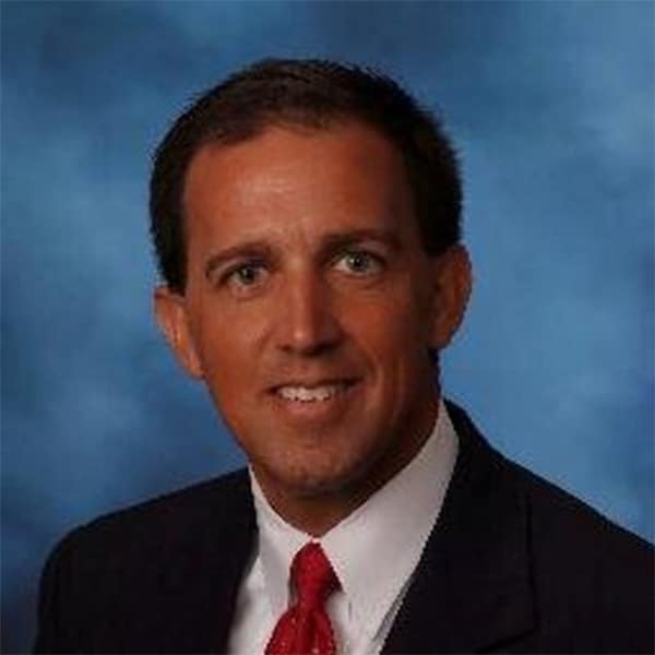 Professional portrait of Greg Rosica, wearing a dark suit, white shirt, and red tie, set against a blue studio background.