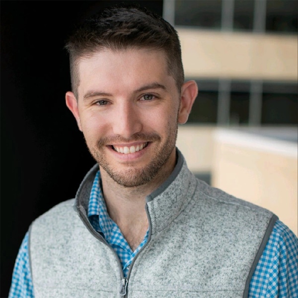 Headshot of Ryan Gilbert, smiling, with short dark hair and trimmed beard, wearing a light gray fleece vest over a blue checkered shirt, with an office building in the background.