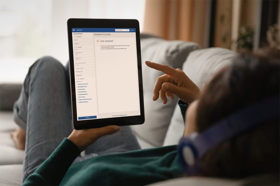Person reclining on a couch wearing headphones, holding a tablet displaying the University of Florida’s online course schedule page with the “Get started!” message visible.