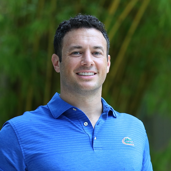 Headshot of Scott Friedman, a man with short curly dark hair and light skin, smiling and wearing a blue polo shirt with the Florida Gators logo, standing outdoors with a blurred green background.