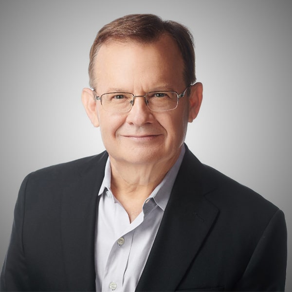 Professional headshot of Steve Rainey, a middle-aged man wearing glasses, a light gray button-up shirt, and a dark blazer, smiling gently against a soft gray background.