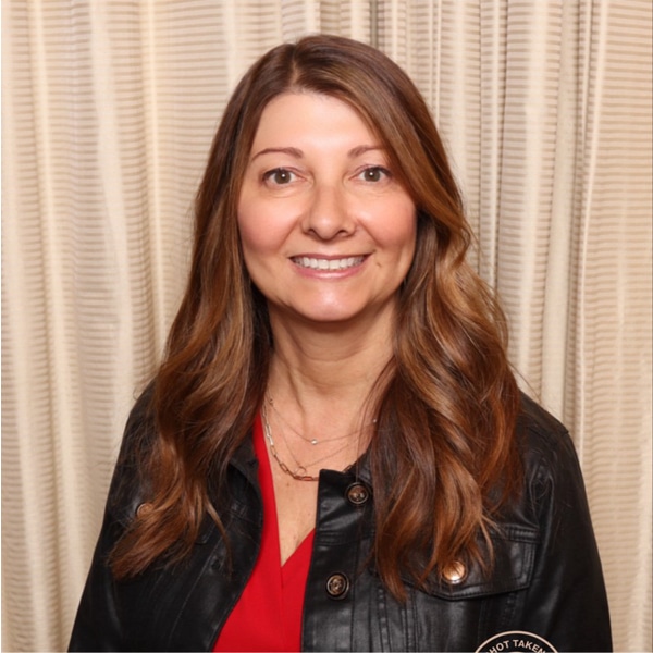 Portrait of Susan Chronister, smiling, with long wavy light brown hair, wearing a black leather jacket over a red blouse, in front of cream-colored curtains.