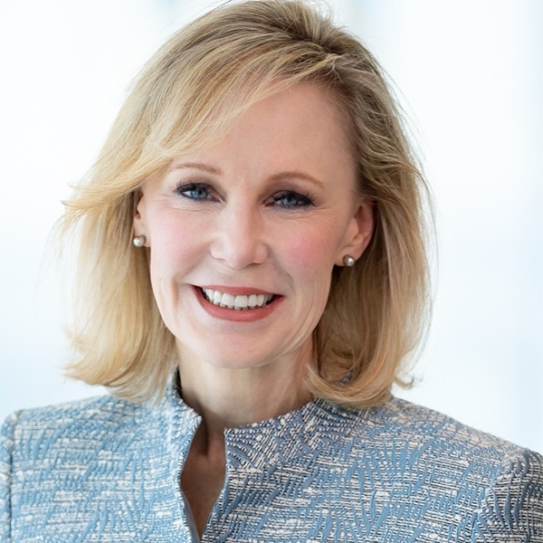 Professional headshot of Suzanne Christensen, a blonde woman with shoulder-length hair, wearing pearl earrings and a patterned light blue jacket, smiling warmly with a bright blurred background.