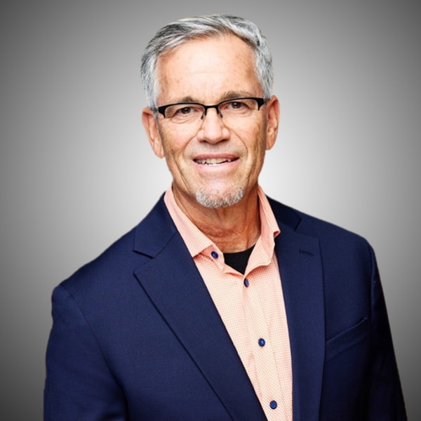 Headshot of Ted McCaffrey, smiling, with short gray hair and glasses, wearing a navy blazer over a salmon button-down shirt, against a gray gradient background.